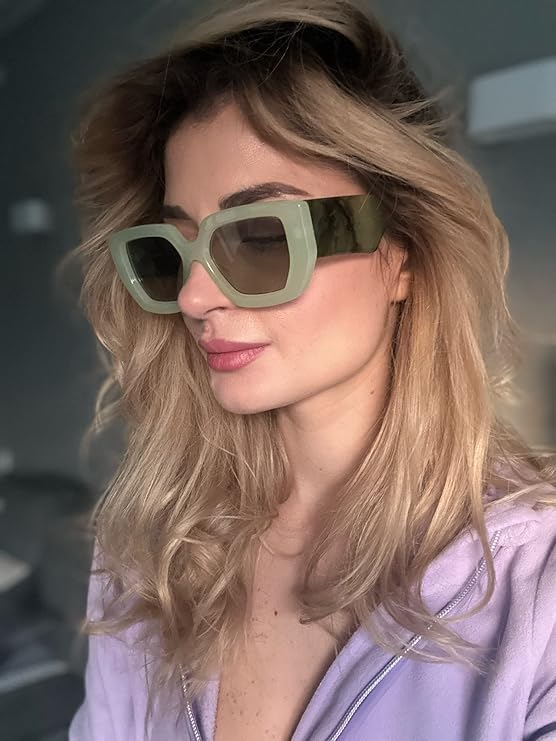 A woman with wavy blonde hair wearing oversized square sunglasses with a green-tinted frame and dark lenses. She is dressed in a light purple zip-up top and has a soft pink lip color. The background is blurred, giving a stylish and modern feel.